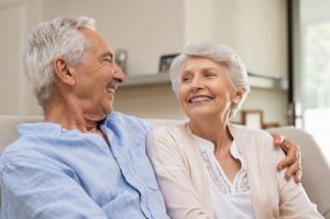 Romantic senior couple laughing while sitting on sofa. Elderly married couple smiling on sofa at home looking at each other. Happy old husband hugging his beautiful wife at home.