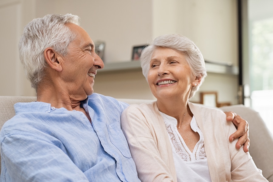 Romantic senior couple laughing while sitting on sofa. Elderly married couple smiling on sofa at home looking at each other. Happy old husband hugging his beautiful wife at home.